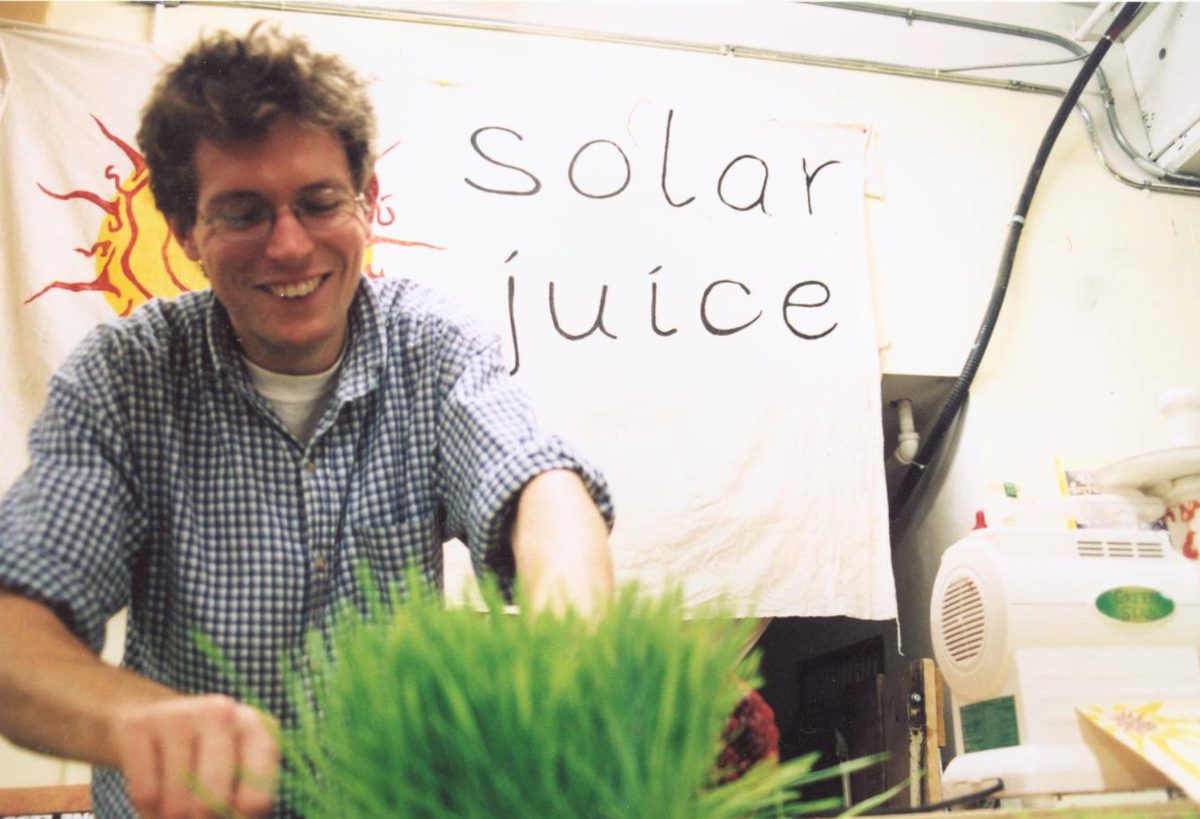 A young Andrew Angus smiling behind his Solar Juice stand in 2001, with a hand-lettered 'solar juice' sign, a painted sun banner, fresh wheatgrass, and a juicer.